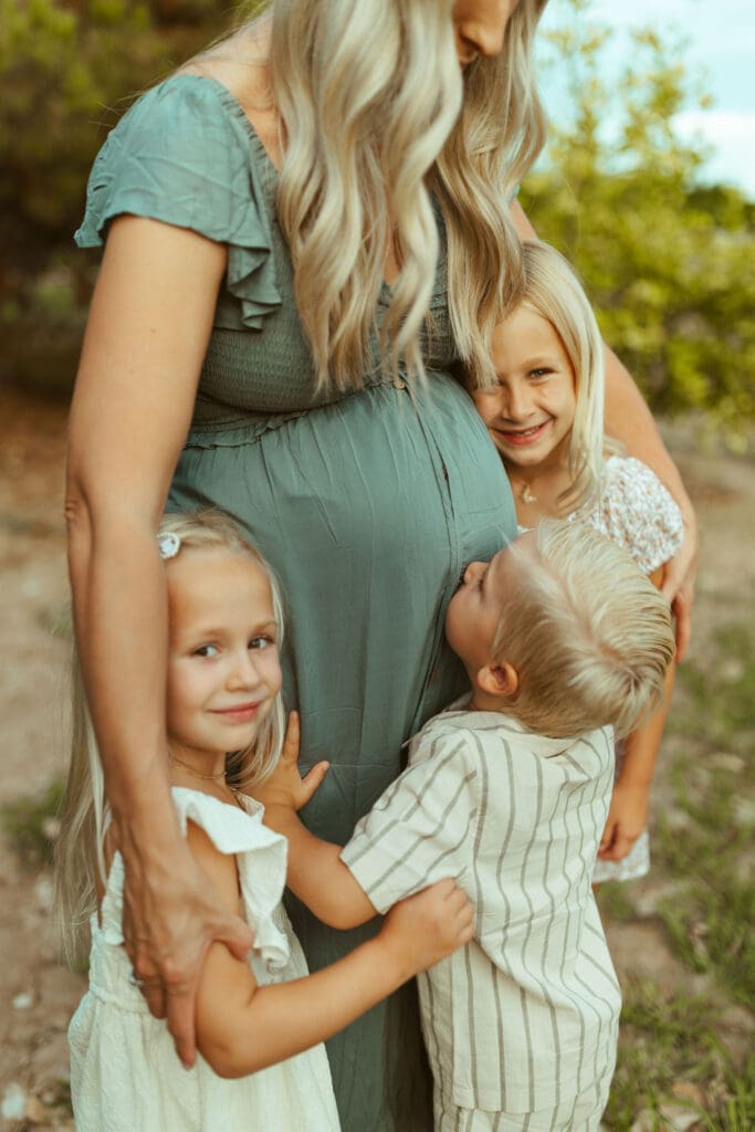 Phoenix maternity photographer capturing three excited siblings hugging their mother's baby bump during family maternity session at Hagen Farms in Queen Creek Arizona