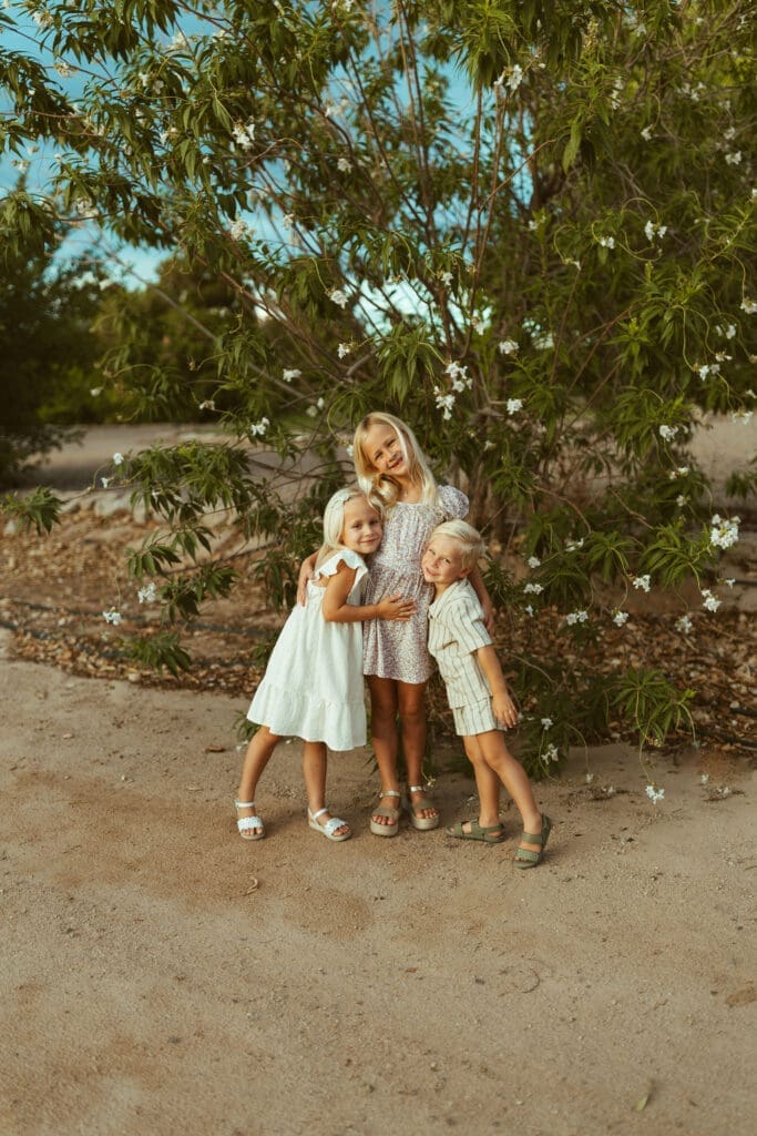 Phoenix maternity photographer capturing expecting mother with three excited children beneath flowering trees at Hagen Farms