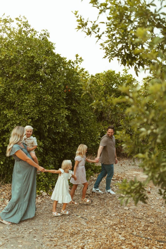 Family walking hand in hand through citrus grove during outdoor maternity photography session near Phoenix Arizona