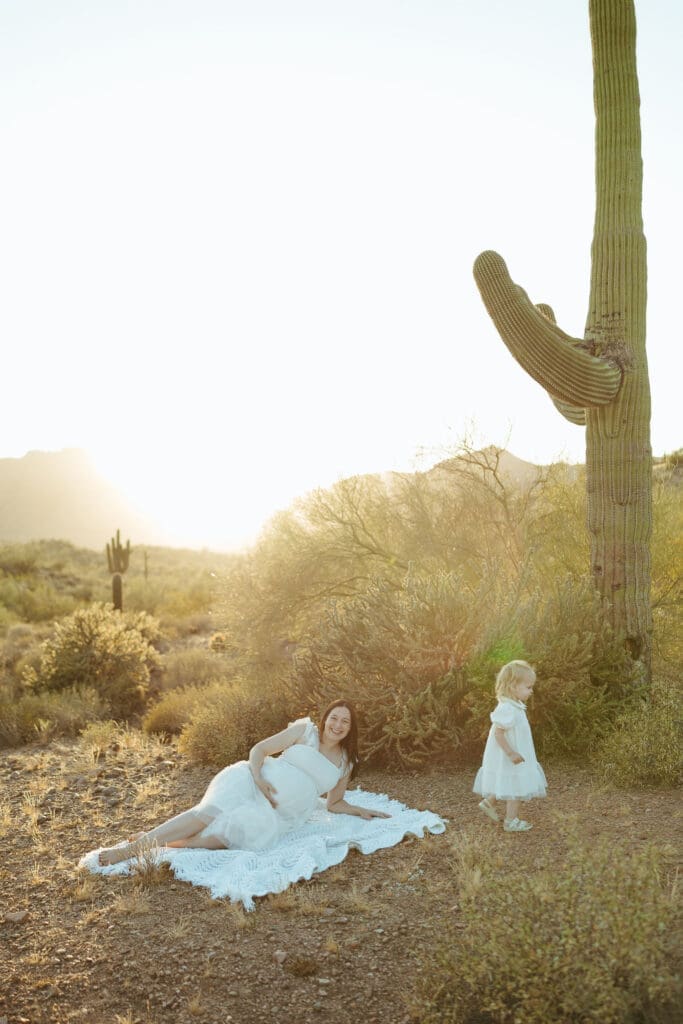 Luxury maternity photography in Arizona featuring mother and toddler on blanket beneath towering saguaro cactus at sunset