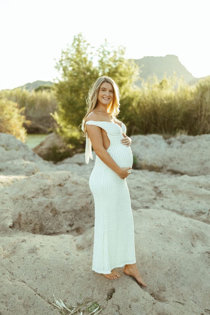 Mother-to-be standing barefoot in the Salt River wearing a fitted white maternity dress during sunset in Mesa Arizona.