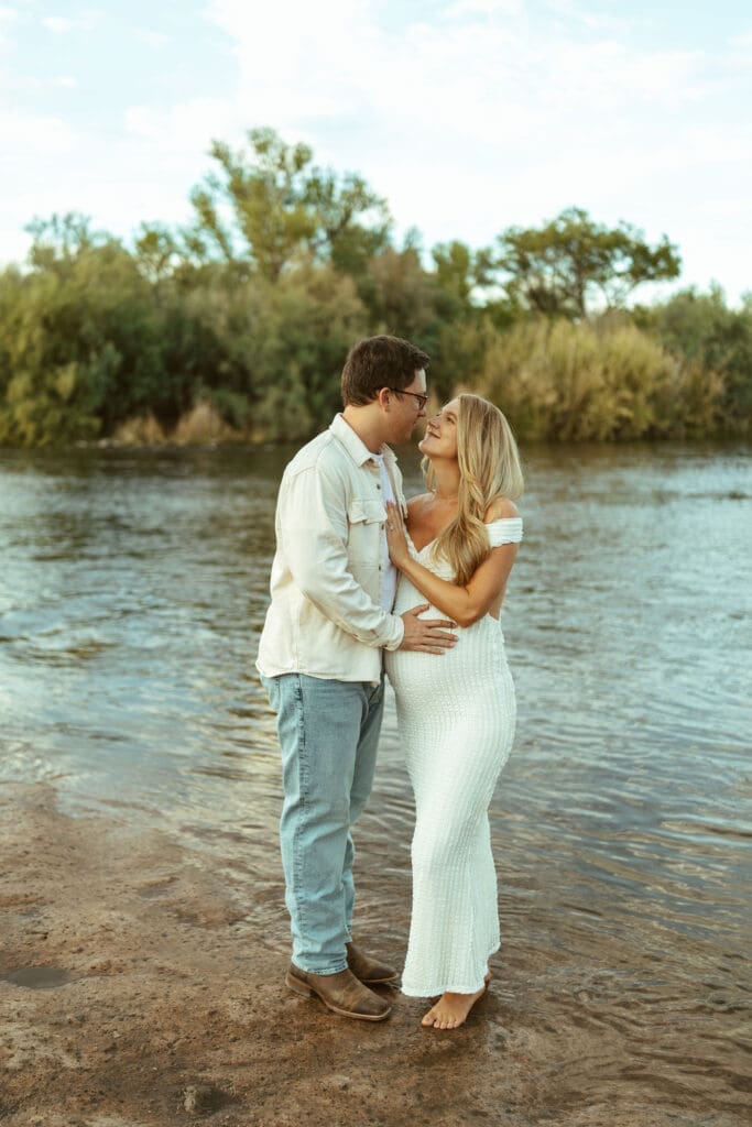 Candid maternity photo of expecting parents smiling together beside the Salt River at golden hour in Arizona.