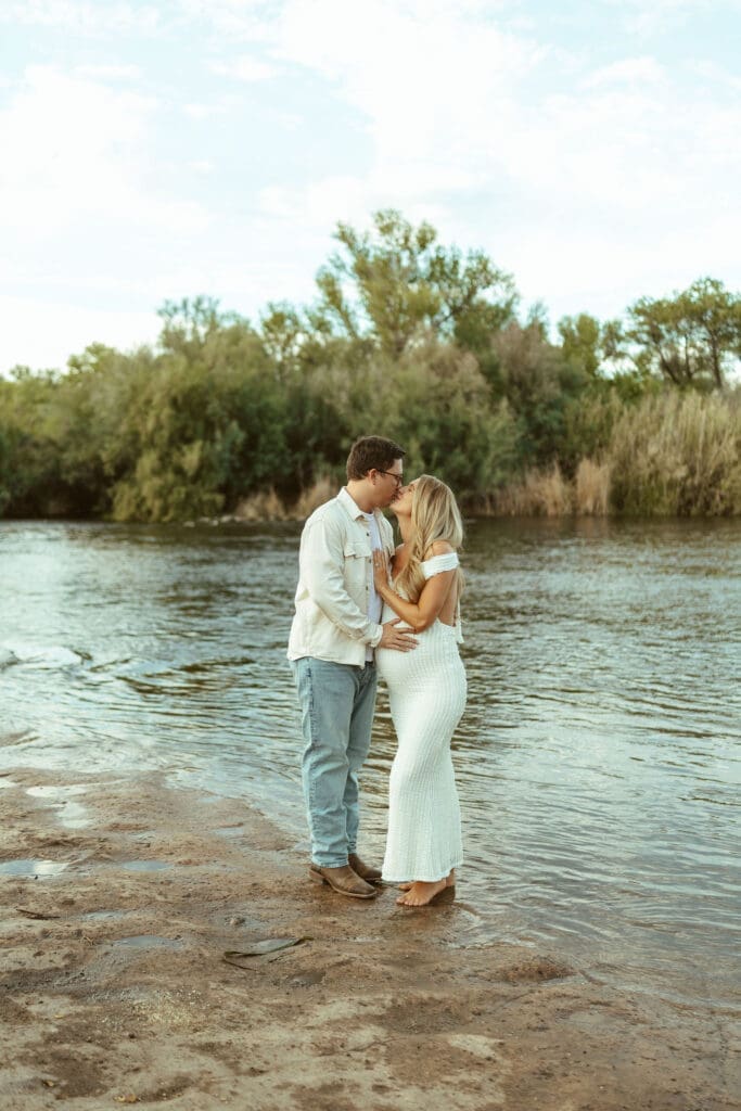 Candid maternity photo of expecting parents smiling together beside the Salt River at golden hour in Arizona.