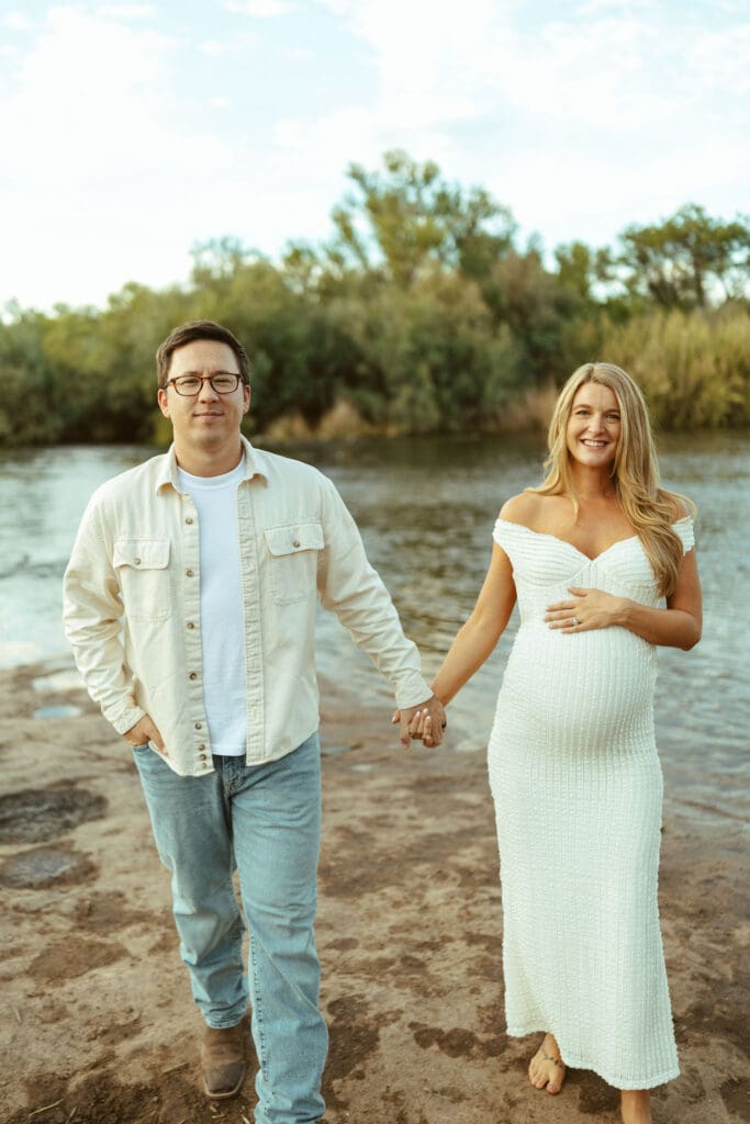 Candid maternity photo of expecting parents smiling together beside the Salt River at golden hour in Arizona.