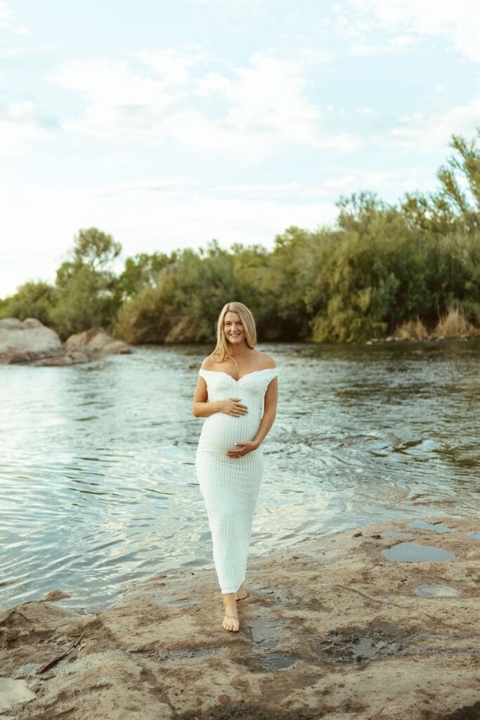 Pregnant woman walking barefoot beside the Salt River in a white fitted maternity dress during sunset in Arizona.