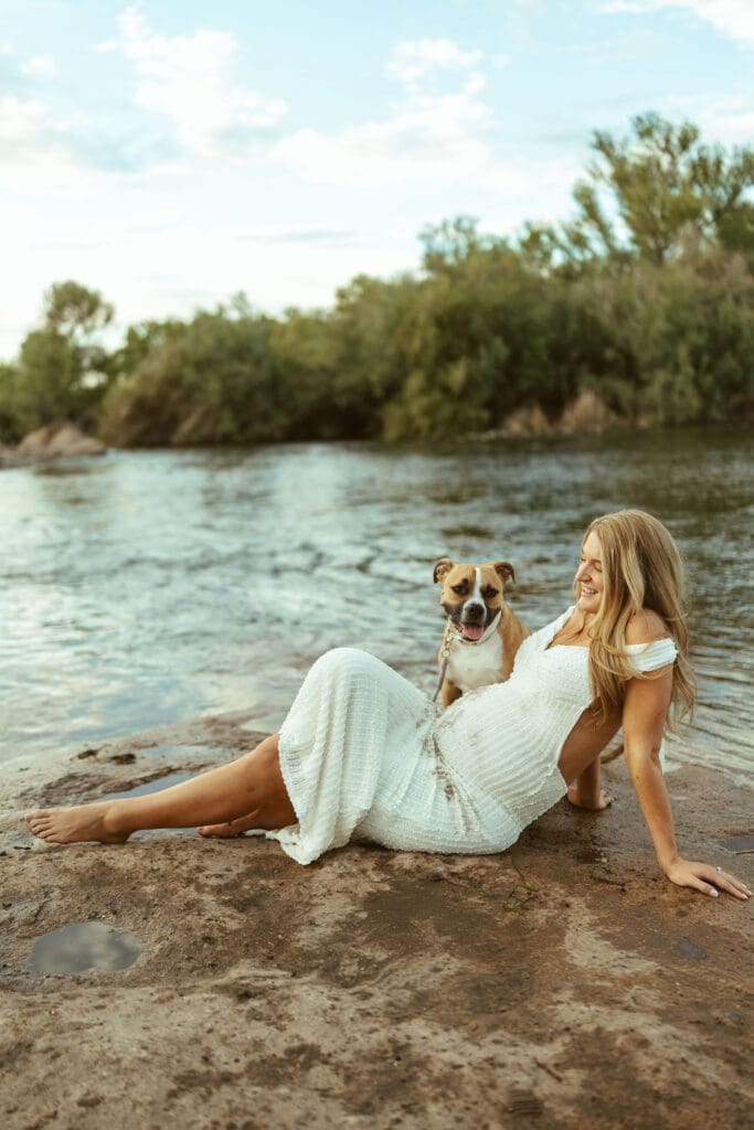 Pregnant woman sitting beside the Salt River with her dog during a sunset maternity session in Mesa Arizona.