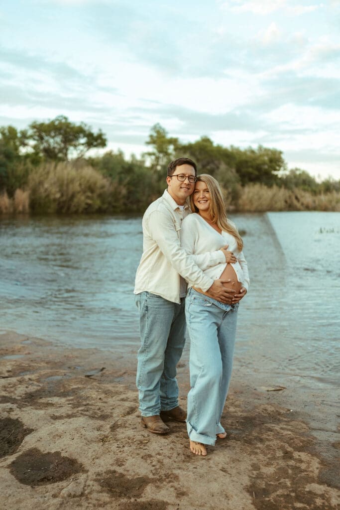 Pregnant couple walking hand in hand beside the Salt River during a golden hour maternity shoot in Arizona.