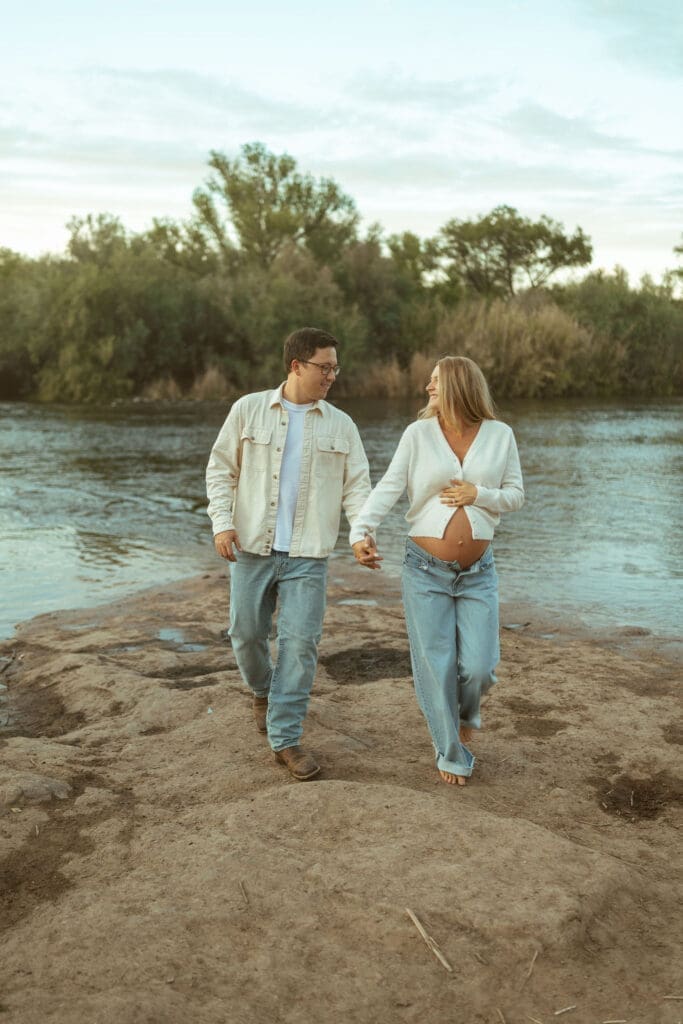 Pregnant couple walking hand in hand beside the Salt River during a golden hour maternity shoot in Arizona.