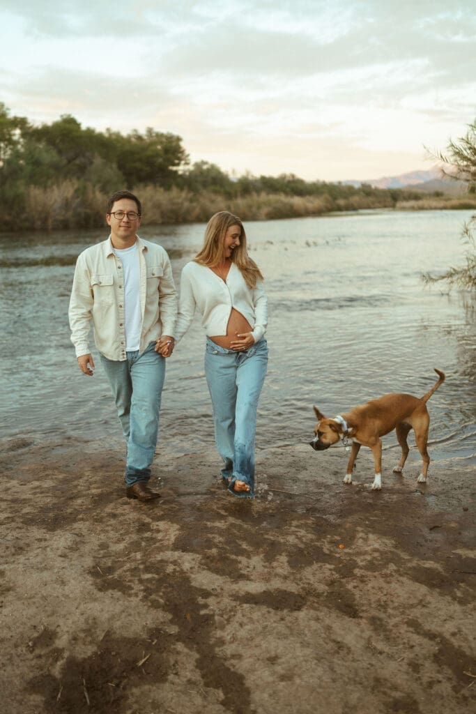 Pregnant couple walking beside the Salt River with family dog during sunset maternity photos.