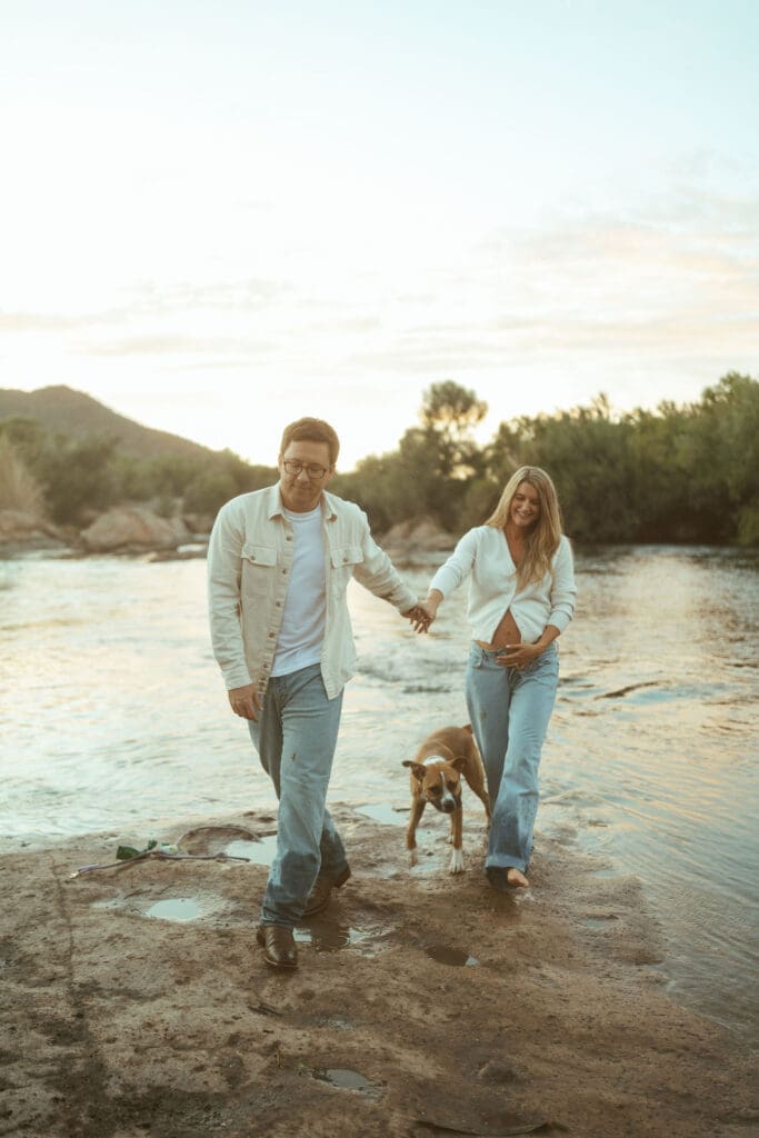 Couple walking hand in hand with their dog beside the Salt River during a golden hour maternity session.