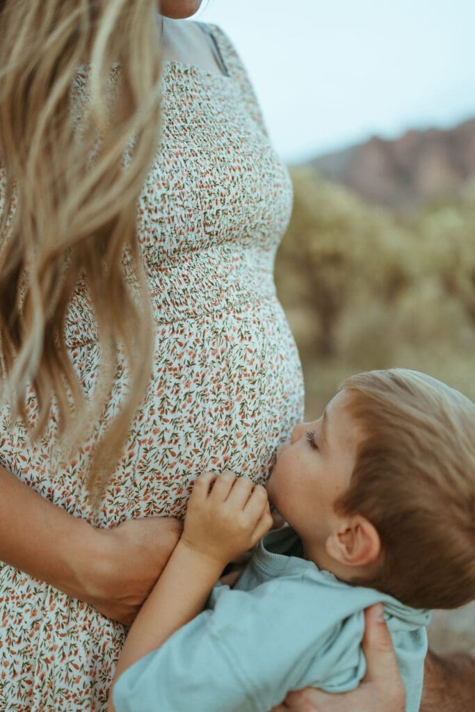 Lifestyle family photography session in Apache Junction Arizona featuring warm sunset light and cactus scenery. Son kissing mothers belly, pregnancy announcement