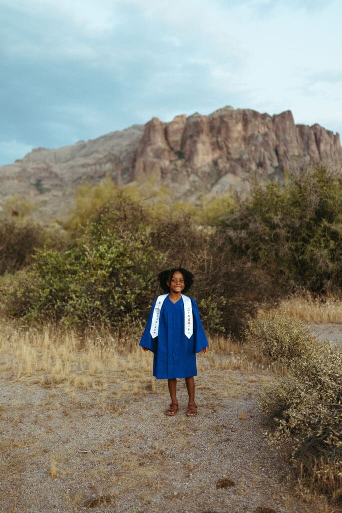 Kindergarten graduate in a blue cap and gown standing in front of the Superstition Mountains