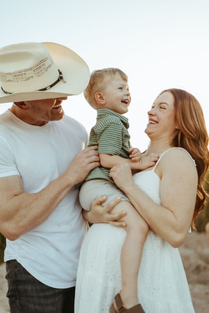 Family maternity session at the Salt River in Mesa, Arizona with parents laughing while holding their toddler at sunset.