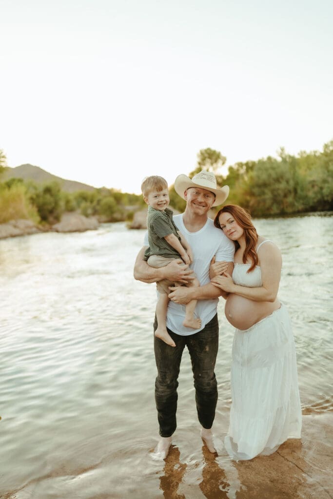Expecting parents and toddler standing together in the Lower Salt River during golden hour maternity photos in Mesa.