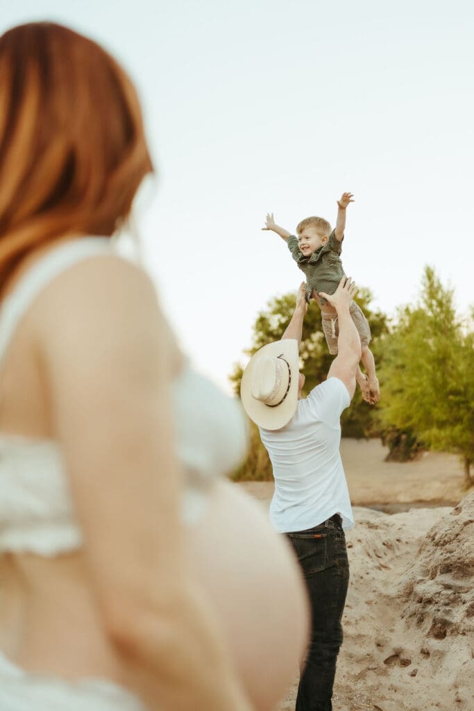 Father tossing his toddler into the air while mom watches during a family maternity session at Phon D Sutton Recreation Area in Mesa, Arizona.
