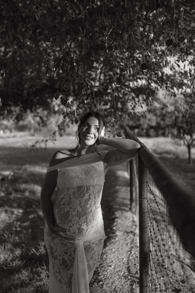 Black and white maternity portrait of an expecting mother standing beneath shaded trees.