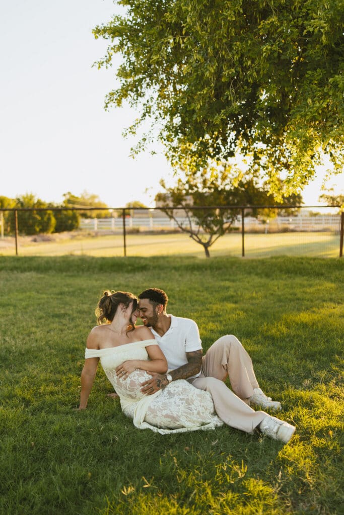 Expecting parents sitting together beneath a large tree during a golden hour maternity photography session.