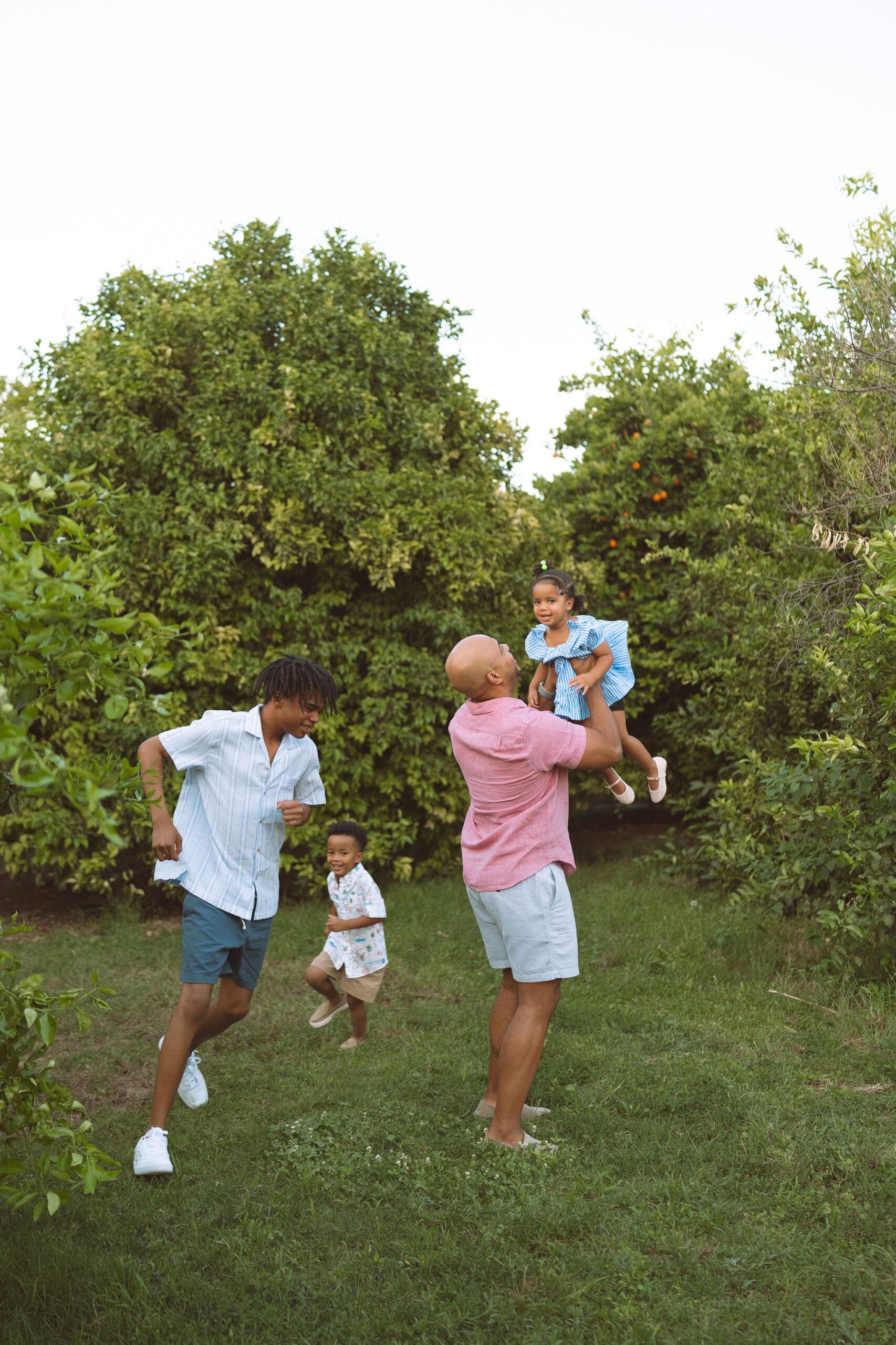 dad in green orchard doing family photos with his three young children. Two sons are running around while dad lifts baby girl and plays with her