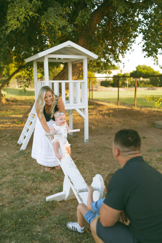 Gilbert family photographer capturing a playful family moment as parents push their daughter on a wooden seesaw during a golden hour outdoor family session.