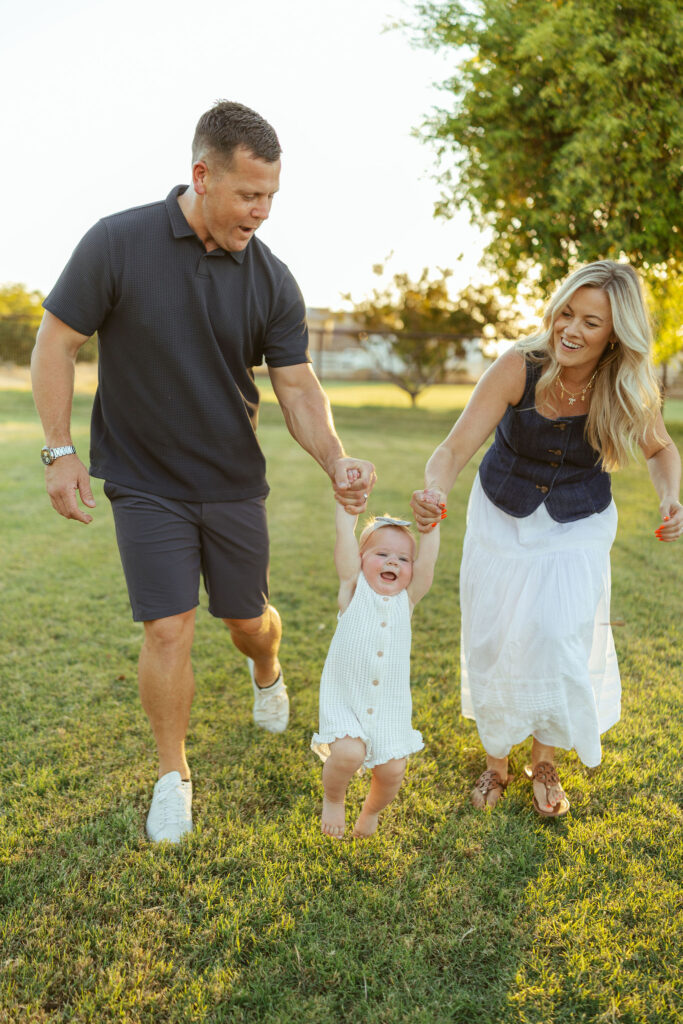 Parents helping their toddler take steps through the grass during an outdoor Gilbert family photography session.