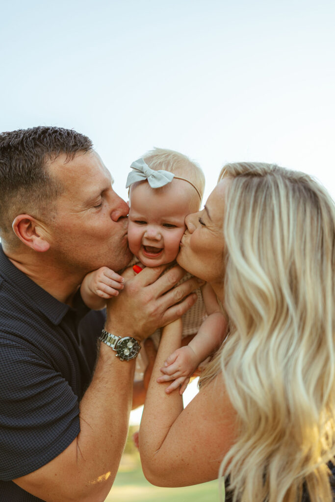 Parents kissing their smiling baby during a golden hour family photography session in Gilbert, Arizona.