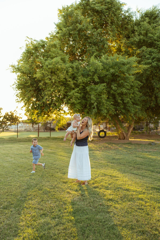 Mother spinning her daughter while her son runs across the grass during a playful Gilbert family photo session.