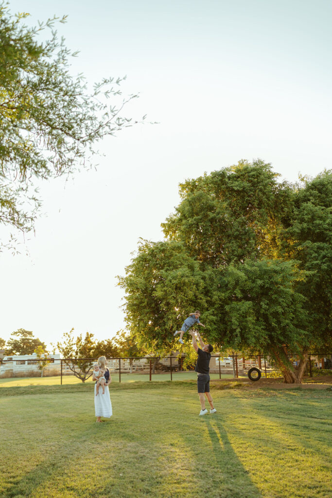 Father lifting his son into the air while mom watches during a joyful golden hour family photography session in Gilbert, Arizona.