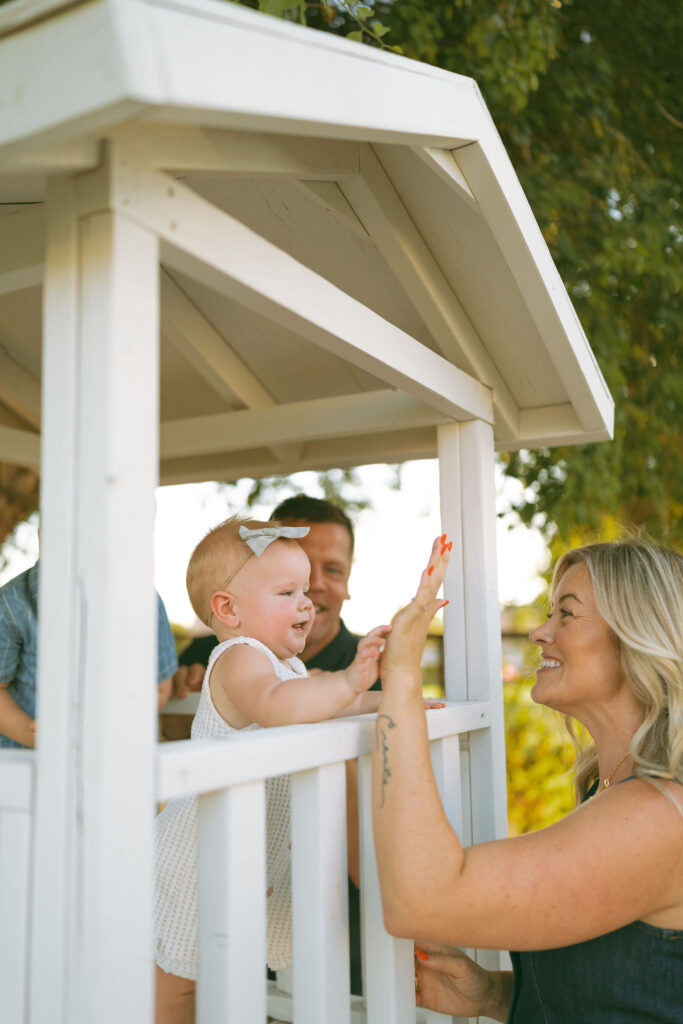 Mother and daughter sharing a high five inside a charming white playhouse during an outdoor Gilbert family photography session.