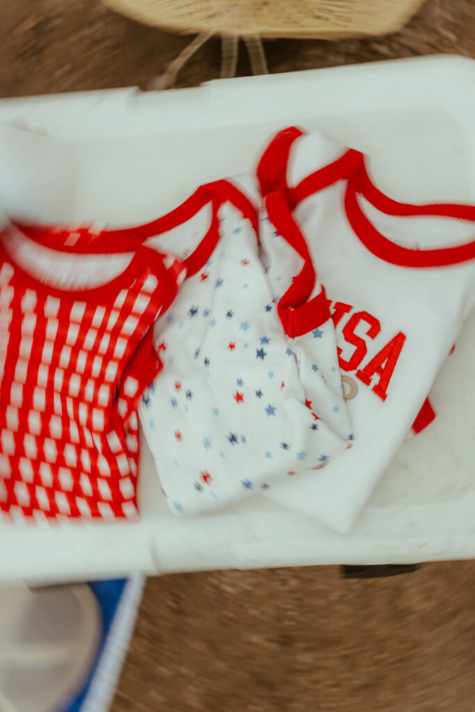Flat lay of patriotic baby outfits, including a red gingham onesie, star-print bloomers, and a USA shirt for a Fourth of July-inspired pregnancy announcement.