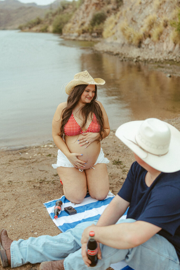 Mesa maternity photographer capturing an expecting mother smiling while sitting on a picnic blanket beside Saguaro Lake during a summer maternity session.