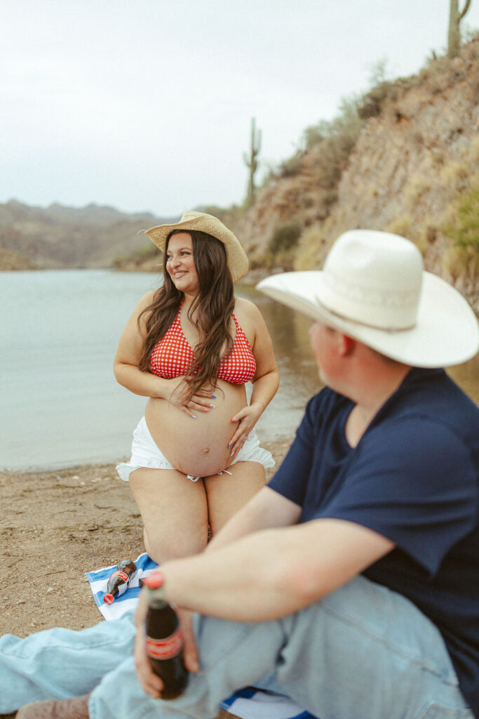 Mesa maternity photographer capturing a couple enjoying a lakeside picnic at Butcher Jones Beach as the expecting mother smiles at her partner while cradling her baby bump.