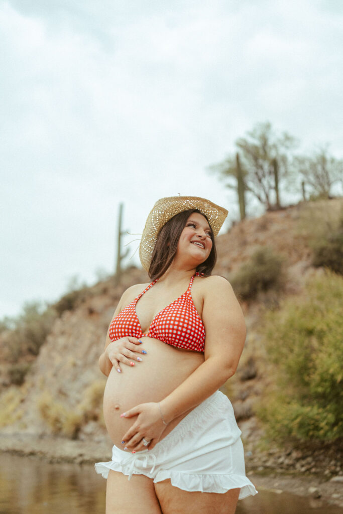 Pregnant mother wearing a red gingham bikini and white shorts during a lakeside maternity session at Butcher Jones Beach in Mesa, Arizona.