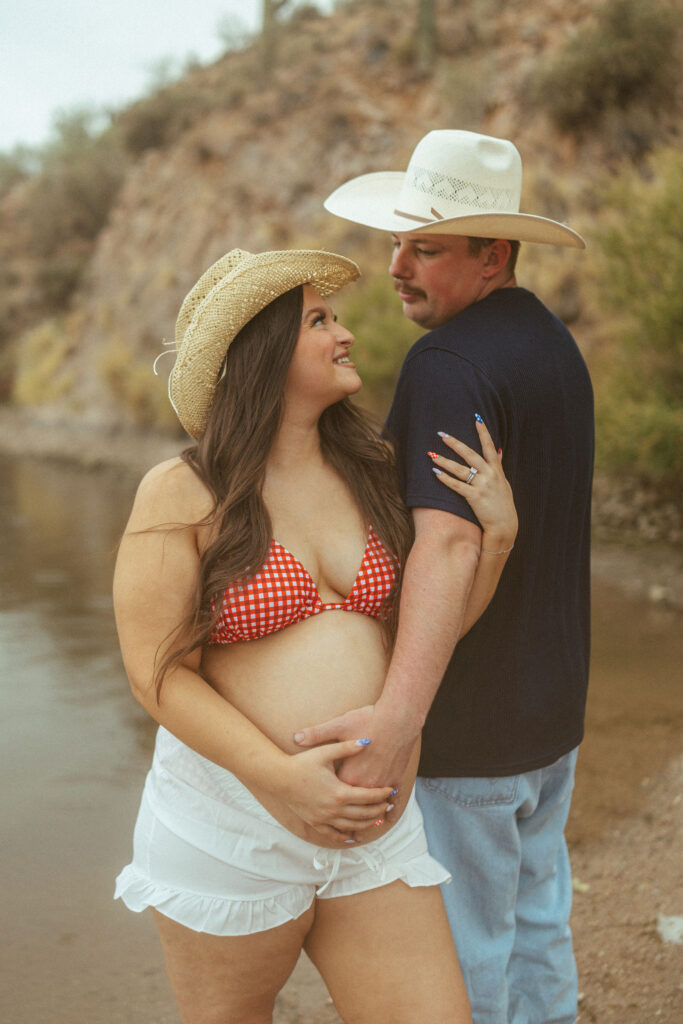 Couple embracing along the shoreline during a romantic maternity session at Butcher Jones Beach with scenic desert mountains in the background.