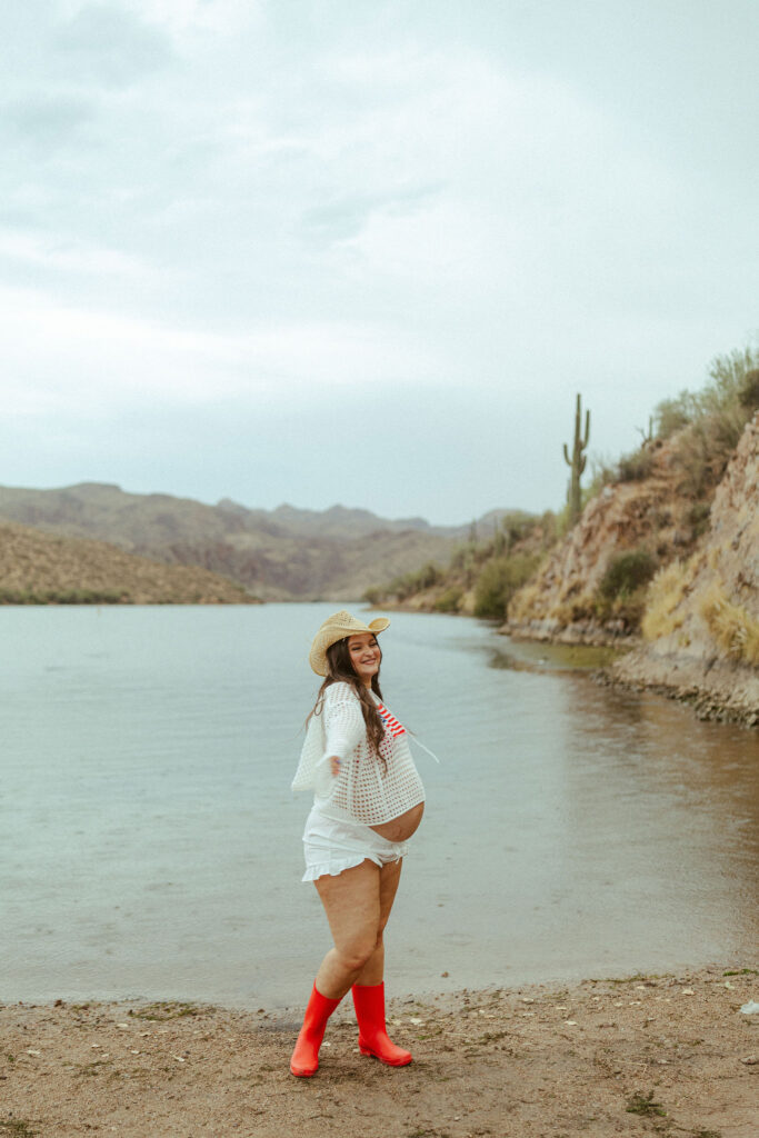 Expecting mother wearing bright red rain boots while standing along the shoreline during a Butcher Jones Beach maternity session in Mesa, Arizona.