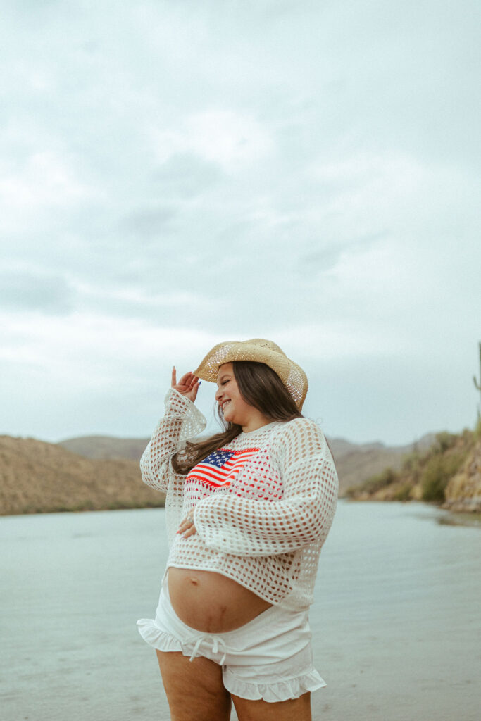 Expecting mother wearing a crochet sweater and cowboy hat while cradling her baby bump during a lakeside maternity session at Saguaro Lake.