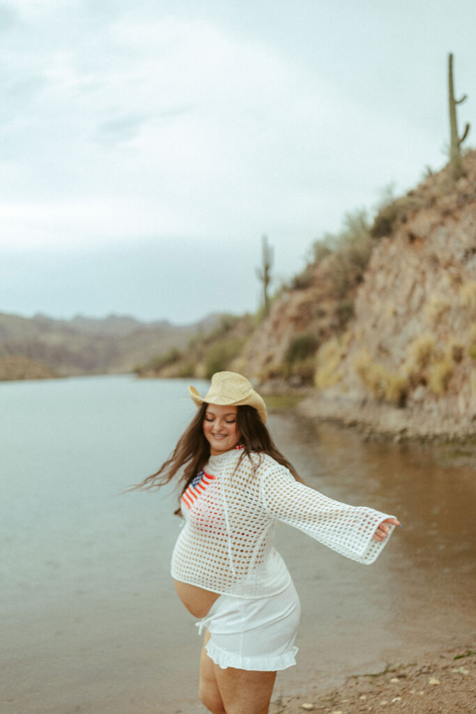 Pregnant mother twirling along the shoreline at Butcher Jones Beach during a summer maternity photography session in Mesa, Arizona.