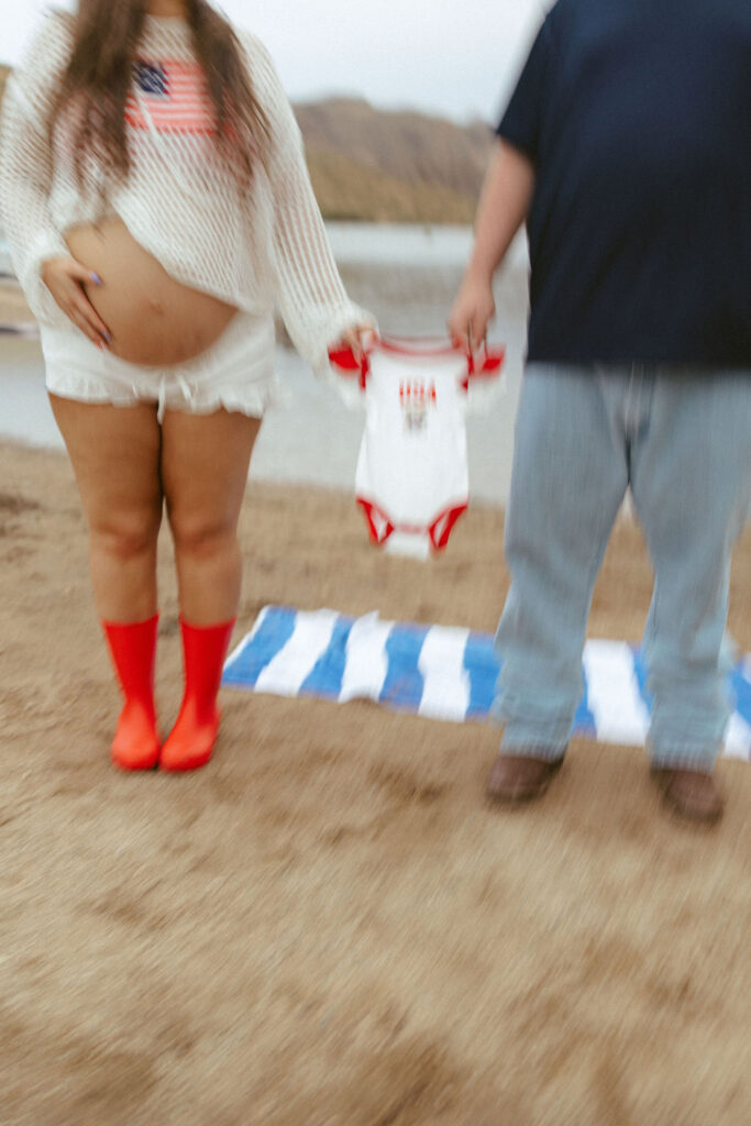 Creative maternity photo of an expecting couple holding a patriotic baby onesie during a pregnancy announcement at Butcher Jones Beach.