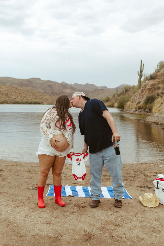 Expecting couple sharing a kiss while holding a patriotic baby onesie during a pregnancy announcement session at Butcher Jones Beach.