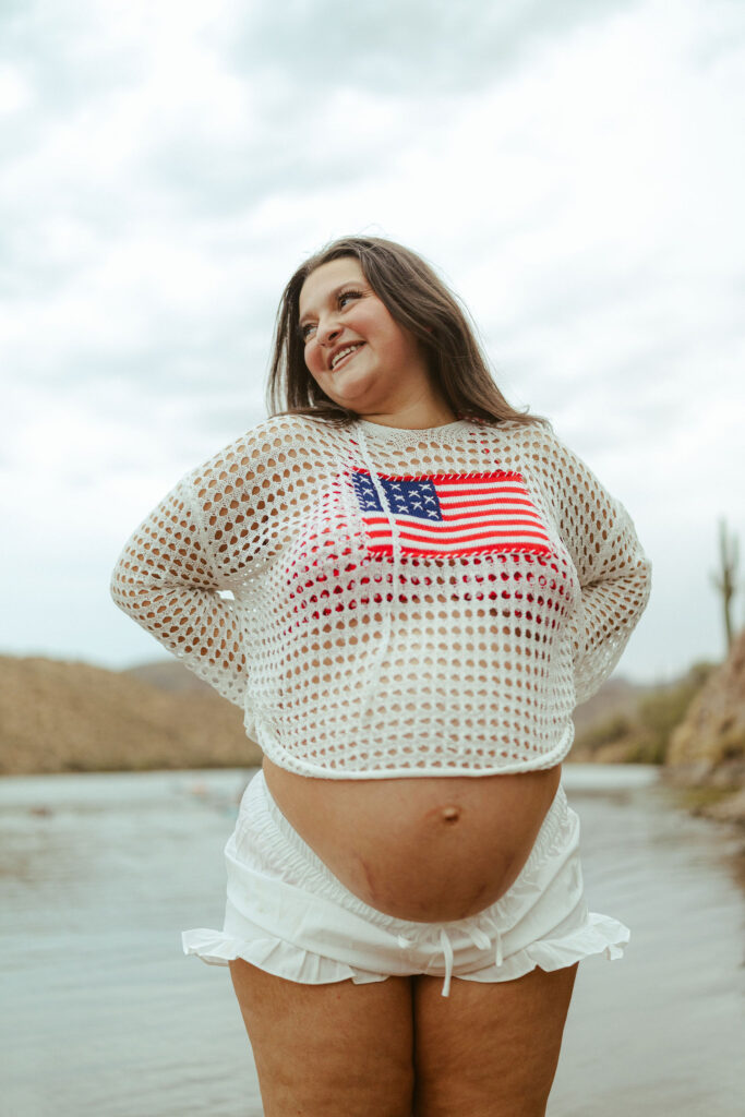 Expecting mother wearing a patriotic crochet sweater while cradling her baby bump during a maternity session at Butcher Jones Beach in Mesa, Arizona.
