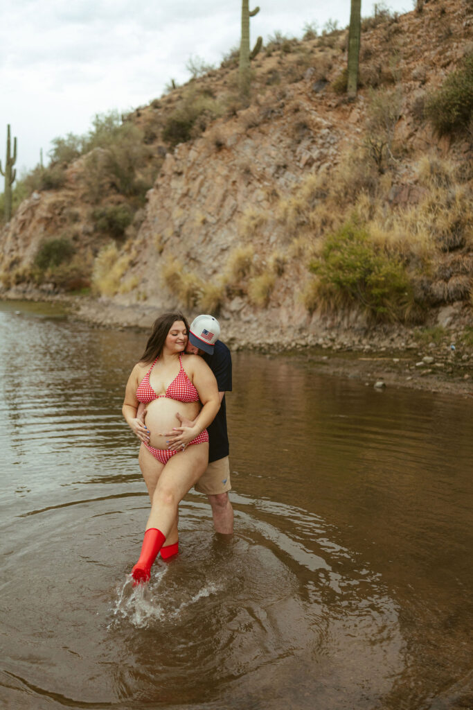 Expecting couple embracing in the shallow water during a romantic maternity session at Butcher Jones Beach with desert cliffs in the background.