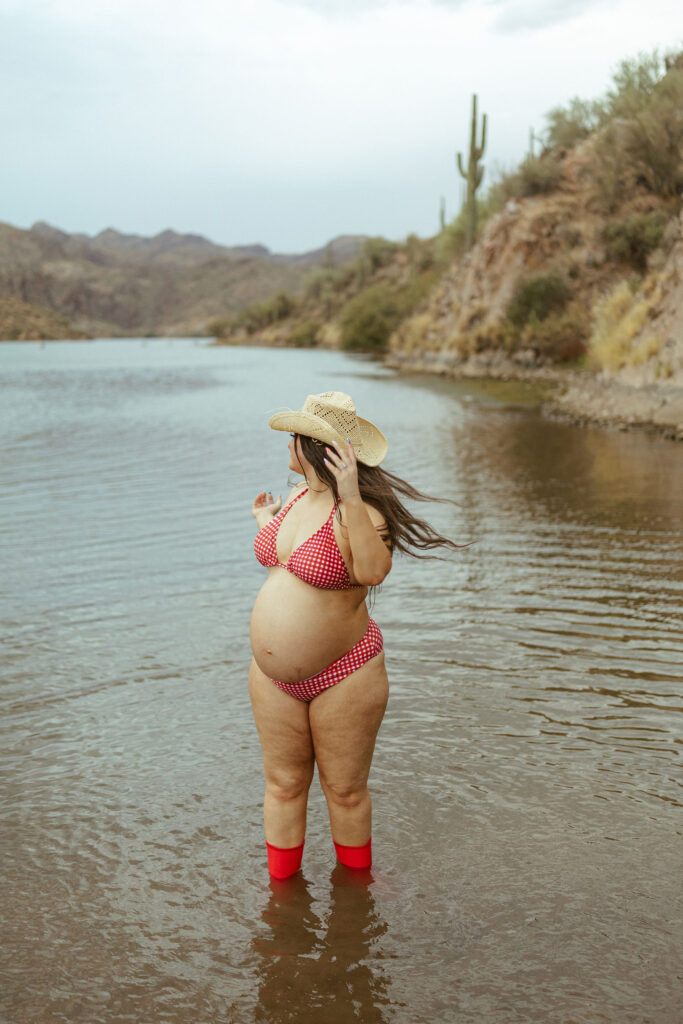 Expecting mother standing in the water wearing a red gingham bikini and cowboy hat during a maternity session at Butcher Jones Beach in Mesa, Arizona.