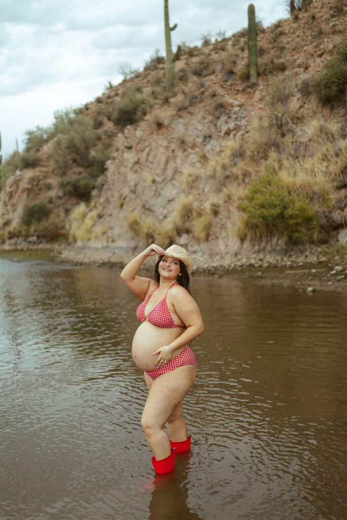 Expecting mother smiling while standing barefoot in the water wearing a red gingham bikini during a lakeside maternity session in Mesa, Arizona.