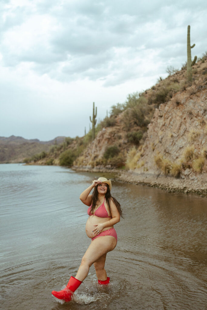 Expecting mother splashing through the water in bright red rain boots during a playful summer maternity session at Saguaro Lake.