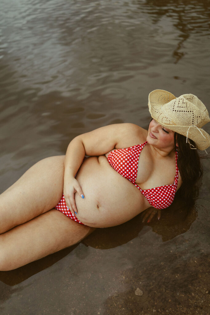 Expecting mother resting in the shallow water while smiling during a summer maternity session at Butcher Jones Beach in Mesa.