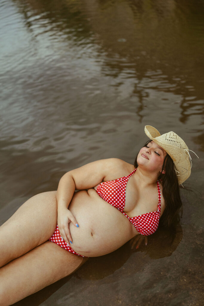 Expecting mother relaxing in the shallow water while wearing a red gingham bikini and cowboy hat during a maternity session at Butcher Jones Beach in Mesa, Arizona.
