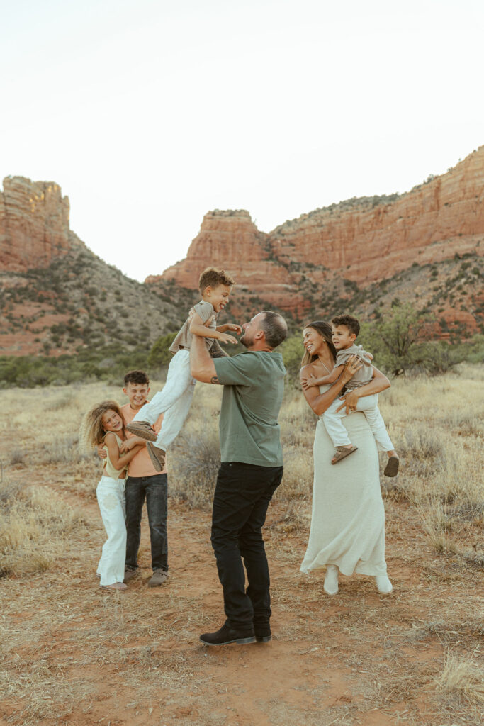 Sedona family photographer capturing a father lifting his son into the air while the rest of the family laughs together in front of Cathedral Rock during a golden hour family session.