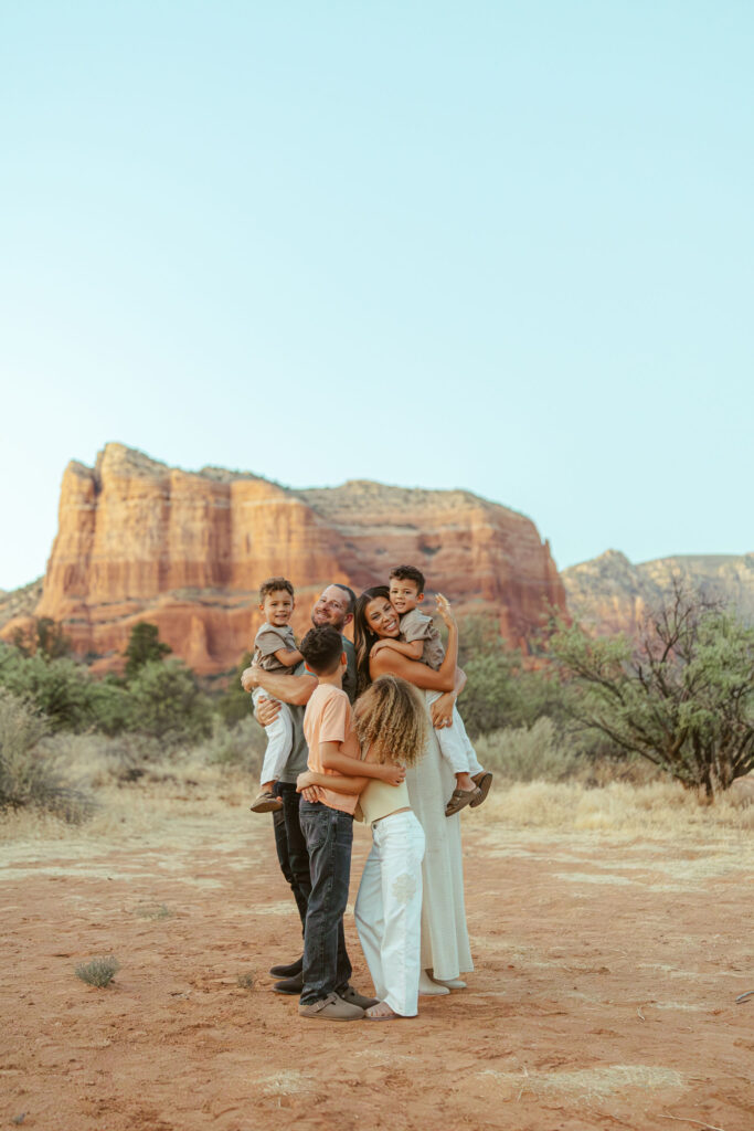 Family of six sharing a joyful group hug during a sunset family photography session in Sedona's iconic red rock landscape.