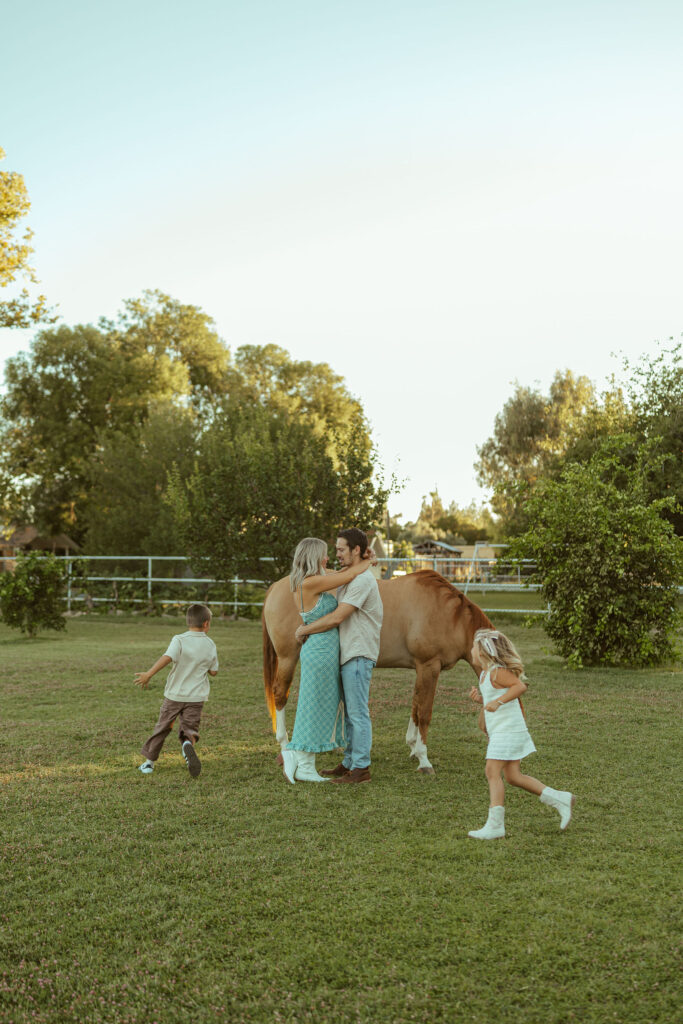 Family photos with horses capturing children running toward their parents during a relaxed pregnancy announcement session in Gilbert, Arizona.