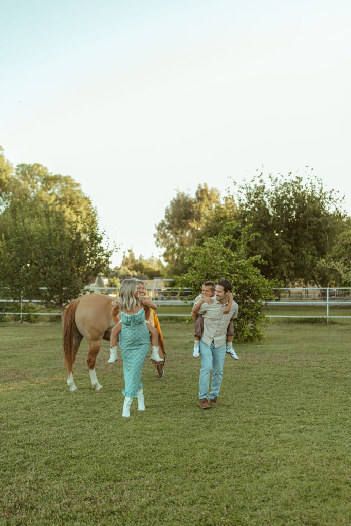 Family farm maternity session featuring parents walking together with their children while their horse grazes in the background in Gilbert.