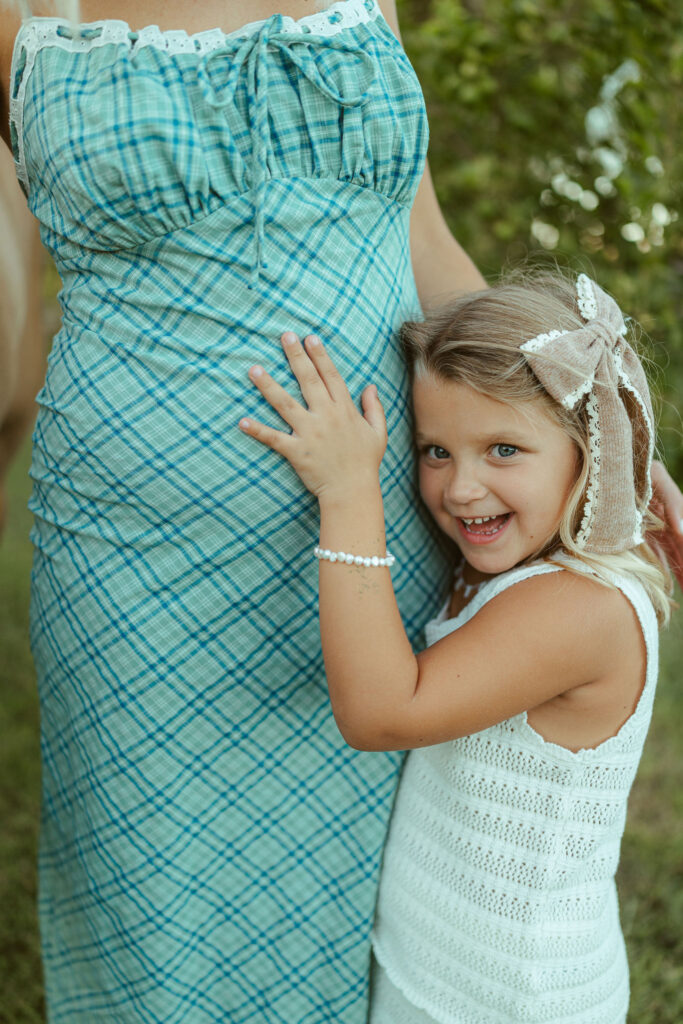 Big sister pregnancy announcement featuring a little girl hugging her pregnant mom's baby bump during an outdoor maternity session in Gilbert, Arizona.